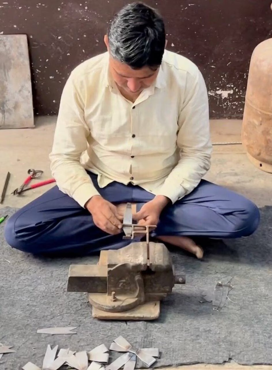 A man sits cross-legged on the floor, working with metal pieces using a bench vise. Tools and metal scraps are scattered around him on a mat, alongside a half-finished Strömshaga Ribbon Hanging Ornament in brass that glints in the light.
