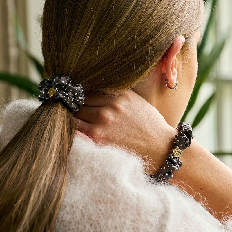 A woman with straight, light brown hair wears a fuzzy white sweater and styles her ponytail with the ByEloise Silk Scrunchie in Polka Dot Mocha with Gold Star. She touches her neck, displaying a matching, snag-free hair accessory on her wrist and wearing a small hoop earring.