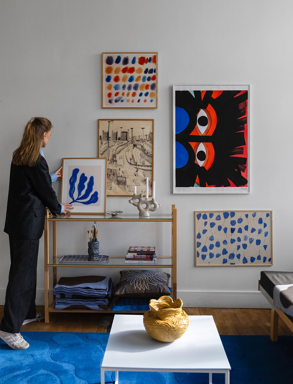 A person arranges the Eyes Print by Tyra von Zweigbergk, 50 x 70 cm (Fine Little Day), above a wooden shelf in a modern living room with a blue rug and a white table topped with a gold decorative bowl.