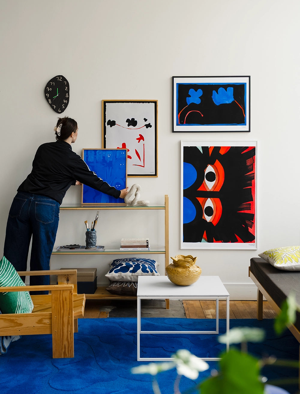 Someone places the Eyes Print by Tyra von Zweigbergk (50 x 70 cm, Fine Little Day) with a blue frame on a wooden shelf in a living room featuring modern art, a blue rug, and minimalist furniture.