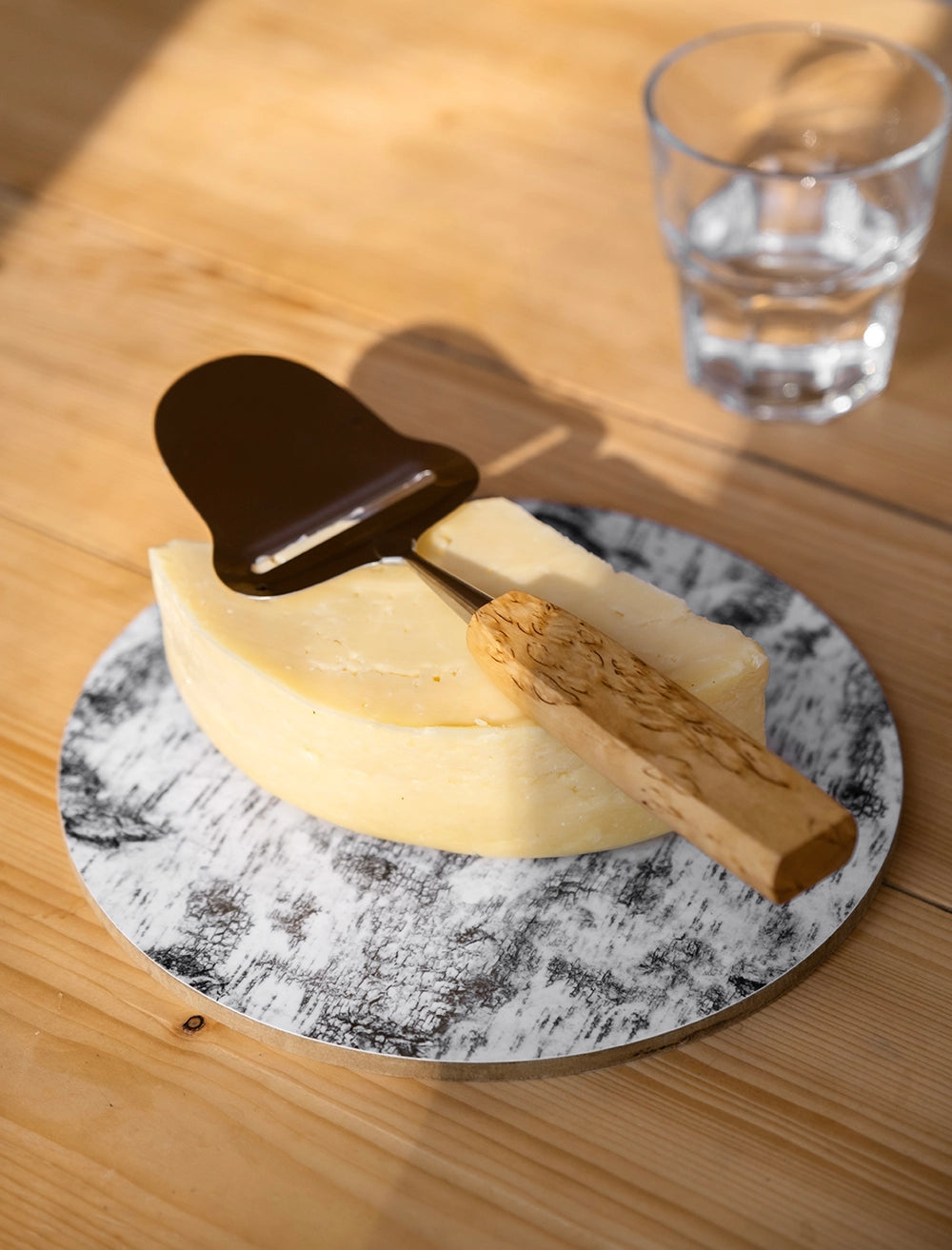 A block of cheese rests on the Fine Little Day Näver Pot Mat (Birch Bark), serving as a stylish table protector, with a cheese slicer placed on top and a glass of water in the background.