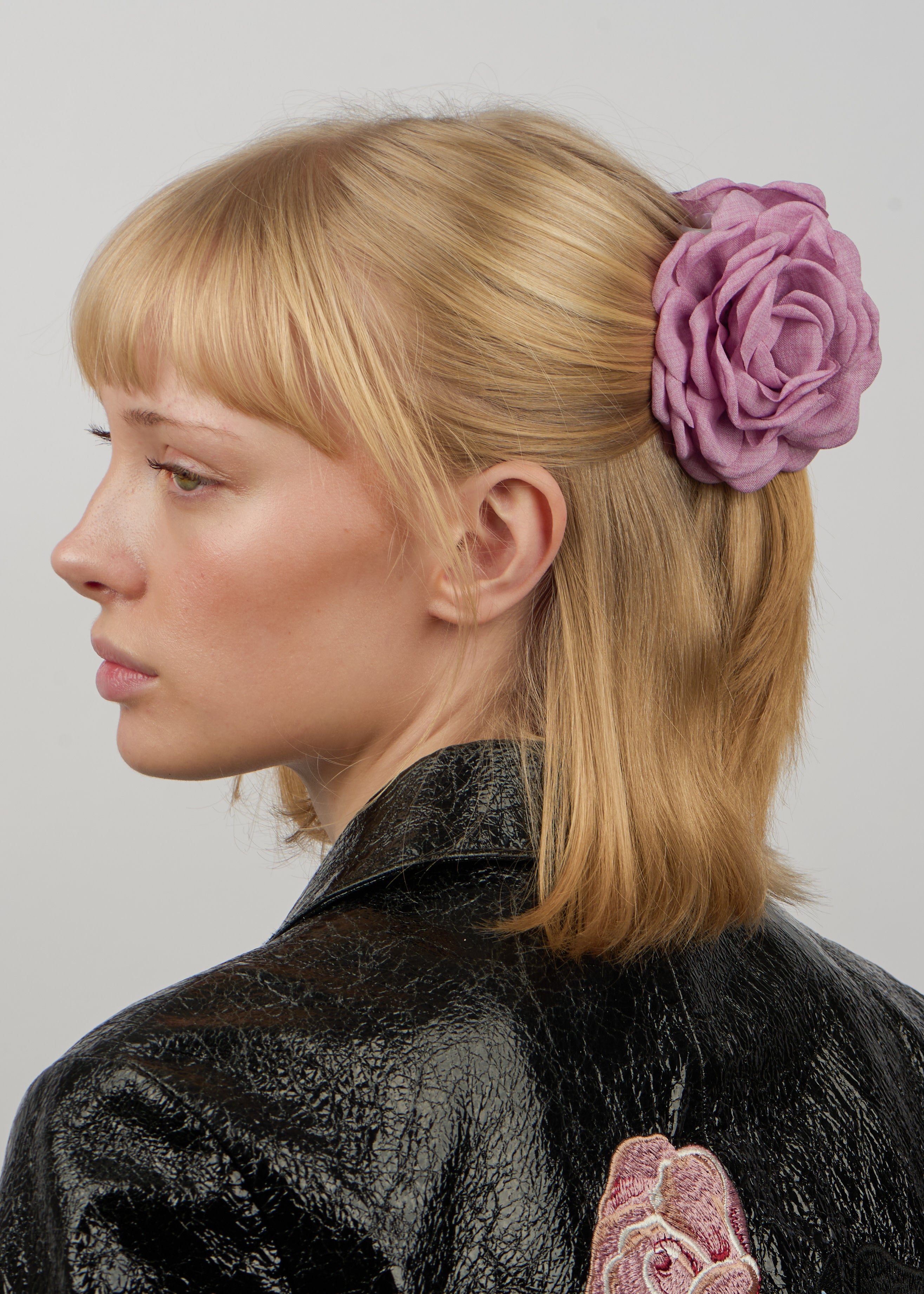 A woman with blonde hair wears the Sui Ava Hair Claw Rosa in Pink Rose, styled in a secure updo, and a black textured jacket with an embroidered dusty rose on the back.