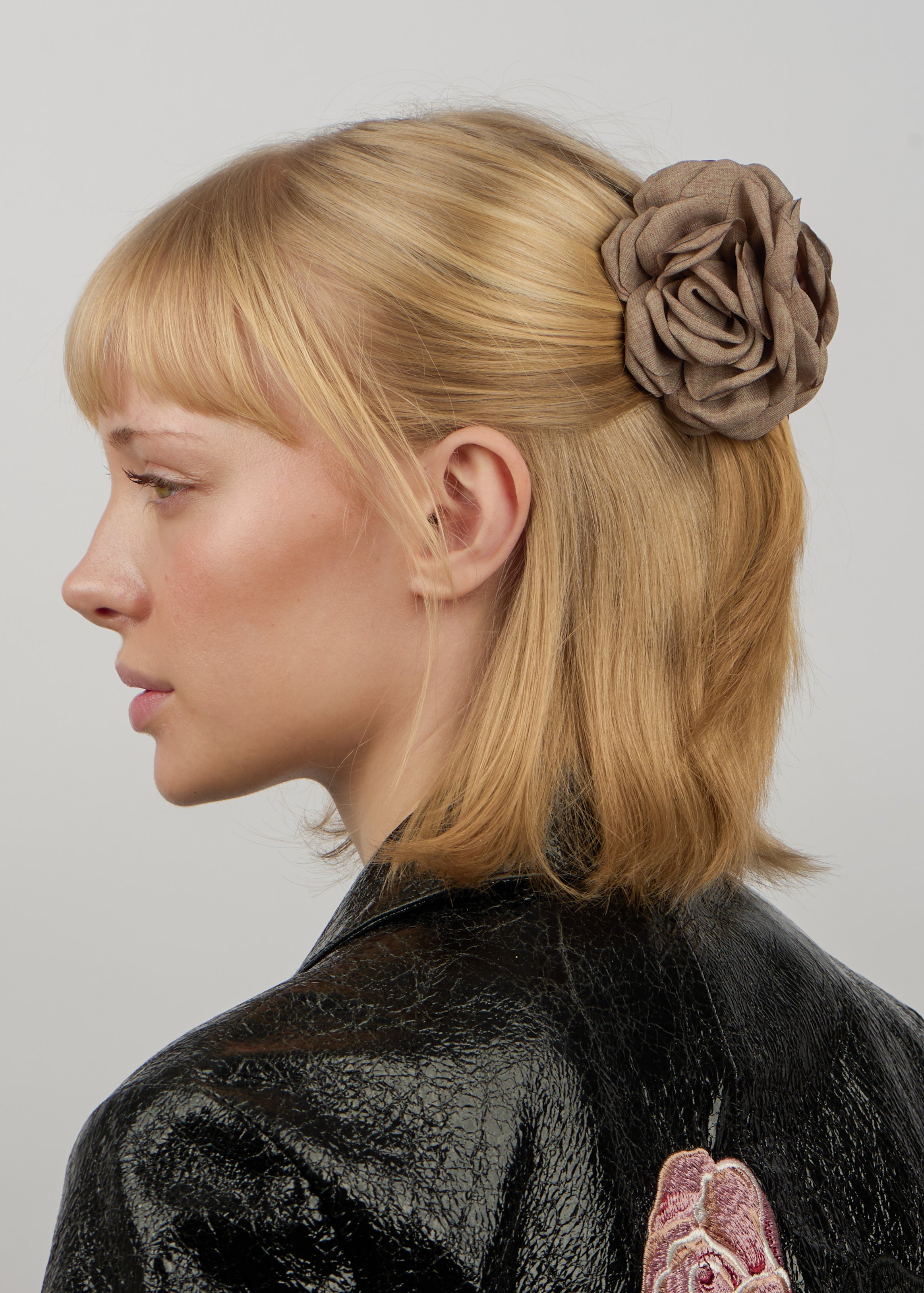 A young woman with blonde hair in a half-up style wears the Sui Ava Hair Claw Rosa in Taupe for a secure hold, shown in profile and paired with a textured black jacket featuring floral embroidery on the back.