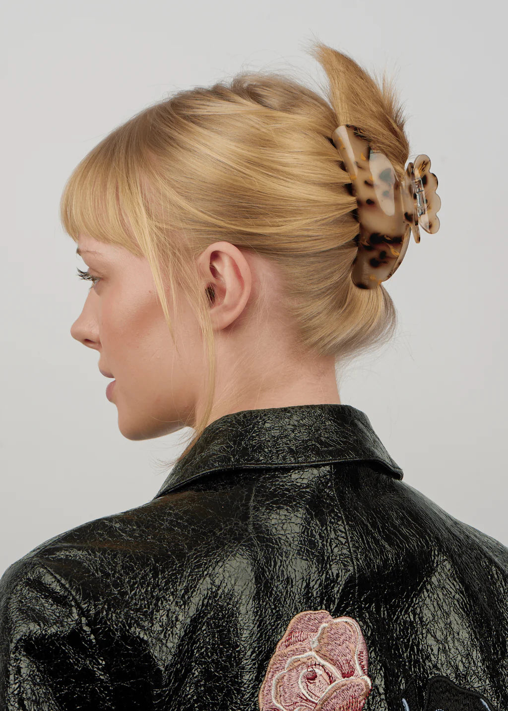 A woman with thick blonde hair is wearing the Sui Ava Helle Pretty Big Hair Claw Clip in Champagne and a textured black jacket with a pink rose embroidery on the back.
