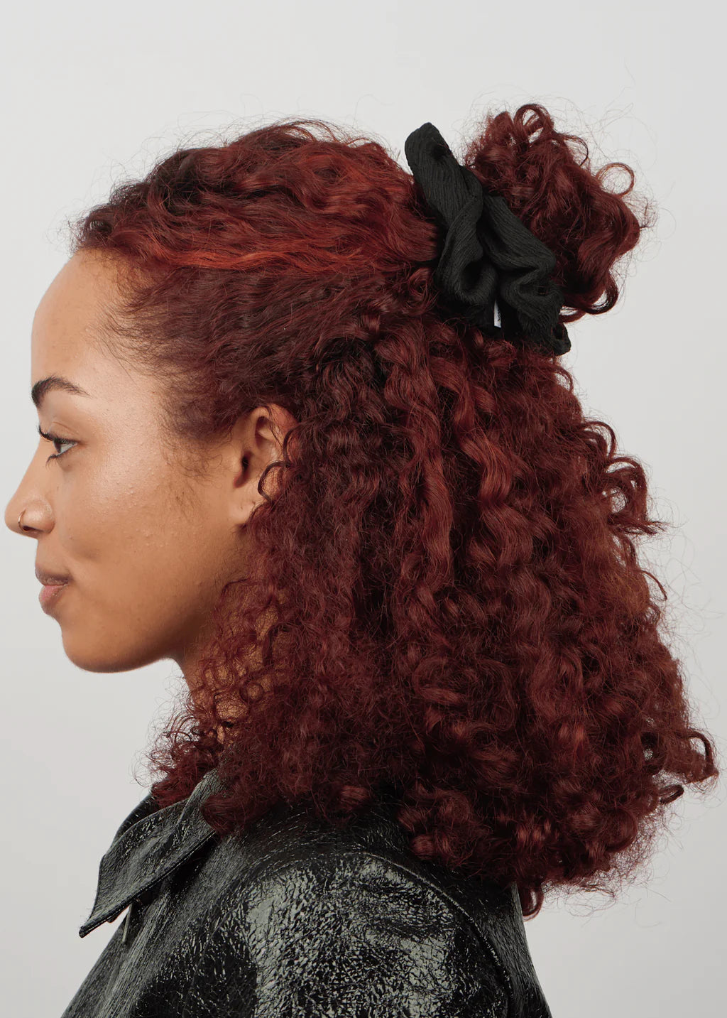 A woman with curly, reddish-brown hair styled in a half-up bun uses the Sui Ava Ea Zig-Zag Scrunchie in Black, paired with a black textured jacket, shown in profile against a plain background.