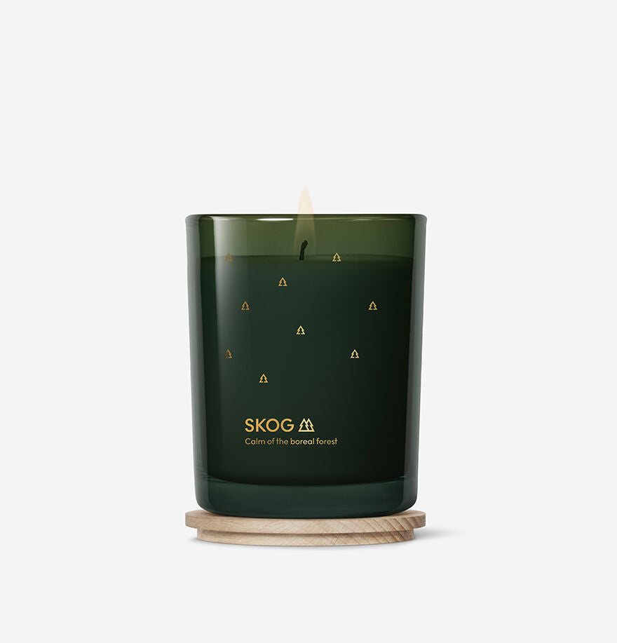 A lit Skandinavisk Seasonal Skog Gold Scented Candle 50h, 200g, made from Swedish rapeseed wax, rests on a round wooden coaster against a plain white background.
