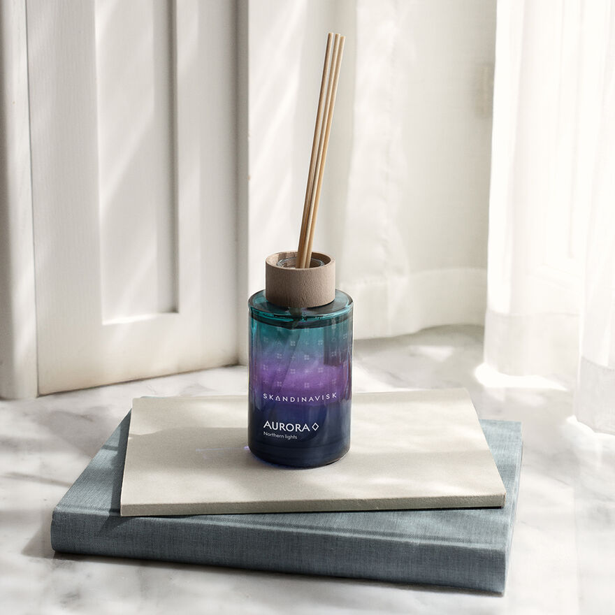 A small, 100 ml Skandinavisk Scent Diffuser Aurora—alcohol-free and finished in blue and purple—rests on two stacked books by a white window, softly illuminated by natural light. Reed sticks diffuse its Aurora scent, with notes of sandalwood and juniper berries that capture the enchanting essence of the aurora borealis.