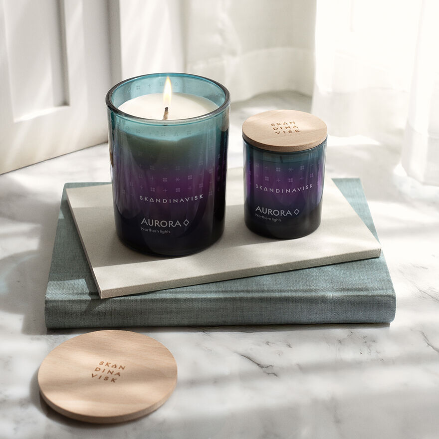 Two Skandinavisk Seasonal Aurora Scented Candles (20h, 65g) crafted from Swedish rapeseed wax rest on stacked books beside a candle lid, all set on a marble surface in soft natural light.