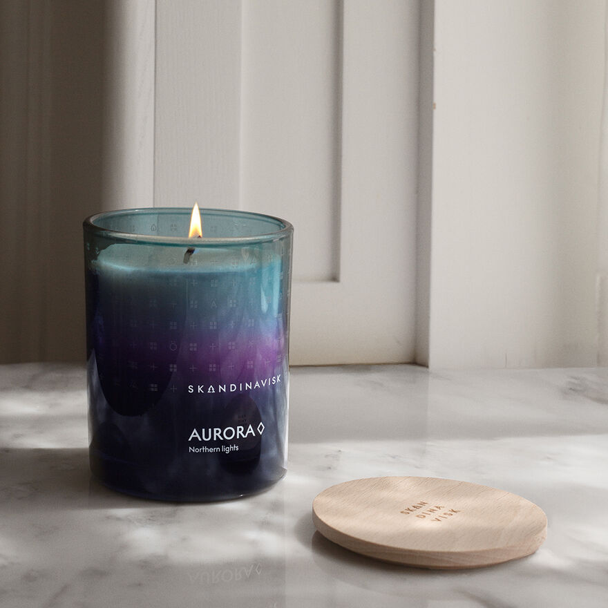 A lit Skandinavisk Seasonal Aurora Scented Candle 50h, 200g, made from Swedish rapeseed wax, sits in a glass jar on a marble surface beside its wooden lid.