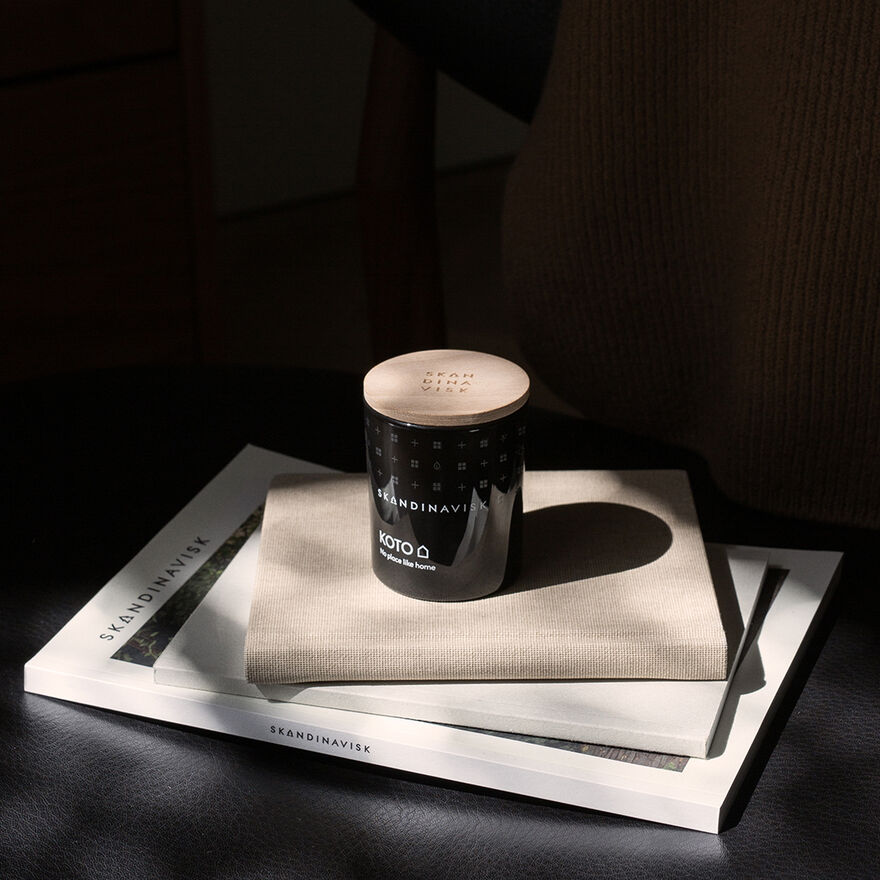 A Skandinavisk Mini Scented Candle Koto 20h, 65g rests on a beige napkin atop two stacked books, one titled SKANDINAVISK, bathed in soft natural light.