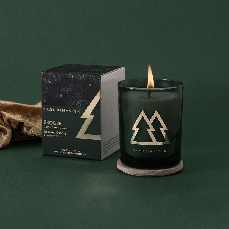 A lit Skandinavisk Seasonal Skog Gold Scented Candle (200g, 50h) glows in a dark green jar with a tree design, pine needle scent filling the air, beside its matching box on a green surface.