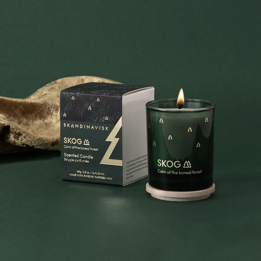 A lit Skandinavisk Seasonal Skog Gold Scented Candle 20h, 65g sits in a green holder on a coaster beside its matching box, both labeled "Calm of the boreal forest," against a dark green background.