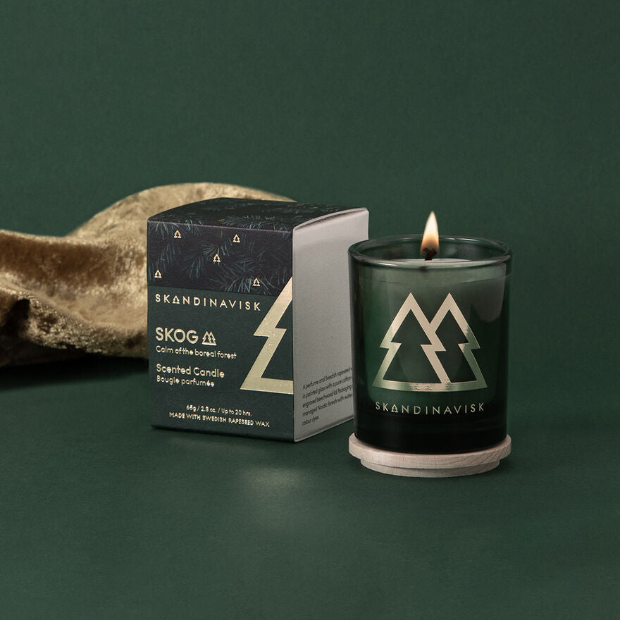 A Skandinavisk Seasonal Skog Gold Scented Candle, 65g, made with Swedish rapeseed wax, sits with its box on a green surface beside a piece of brown fabric.