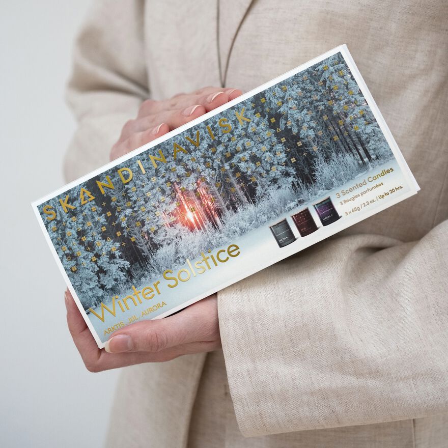 A person in a beige outfit holds the Skandinavisk Scented Candle Winter Solstice Mini Giftset w. Arktis, Jul & Aurora, featuring three mini Swedish rapeseed wax candles with winter forest designs on the packaging.