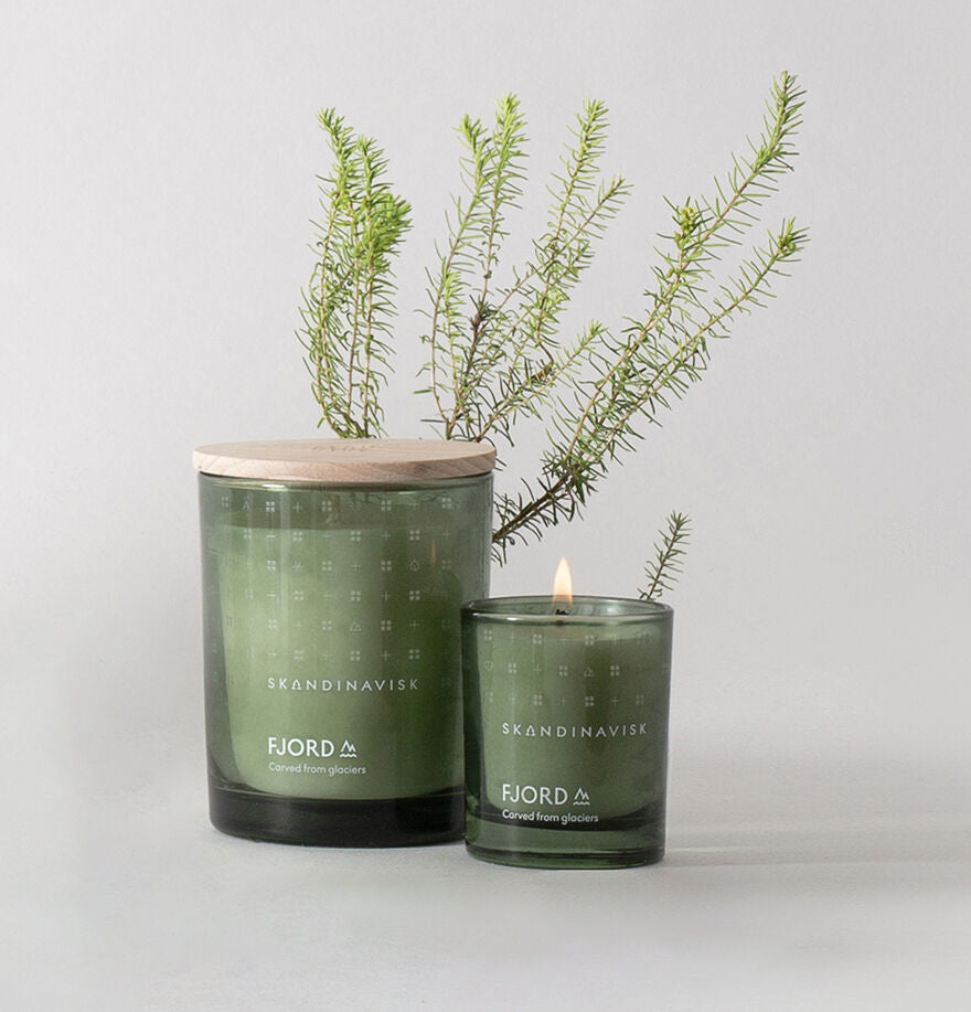 Two green Skandinavisk candles—a large FJORD with engraved beechwood lid and a lit Mini Scented Candle Fjord 20h, 65g—are displayed before green foliage on a light gray background.