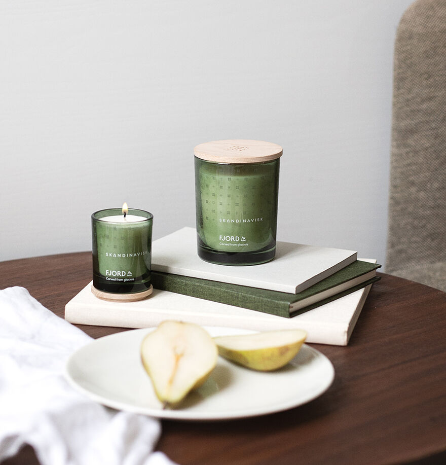 Two Skandinavisk Mini Scented Candle Fjord 20h, 65g candles—one lit, one with an engraved beechwood lid—sit on stacked books next to a plate with sliced pear, all arranged on a wooden table.