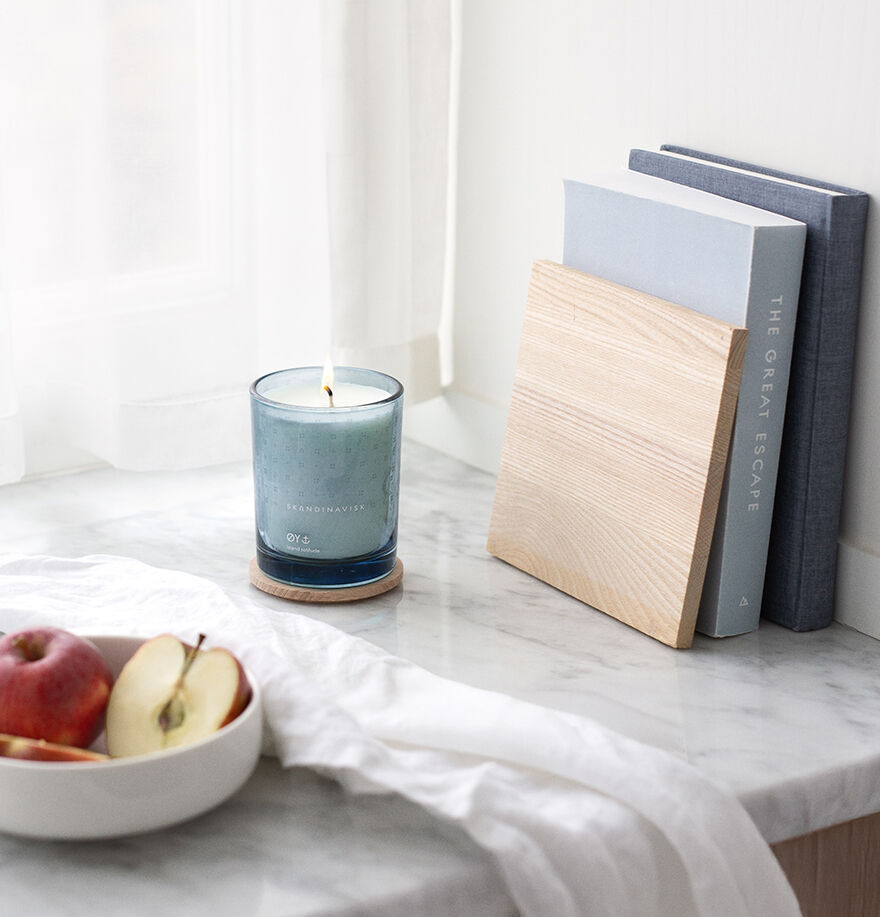 A lit Skandinavisk Scented Candle Øy, 200 g sits on a marble surface beside a bowl of apples and a wooden bookend with two books by a sunlit window draped in sheer white curtains.