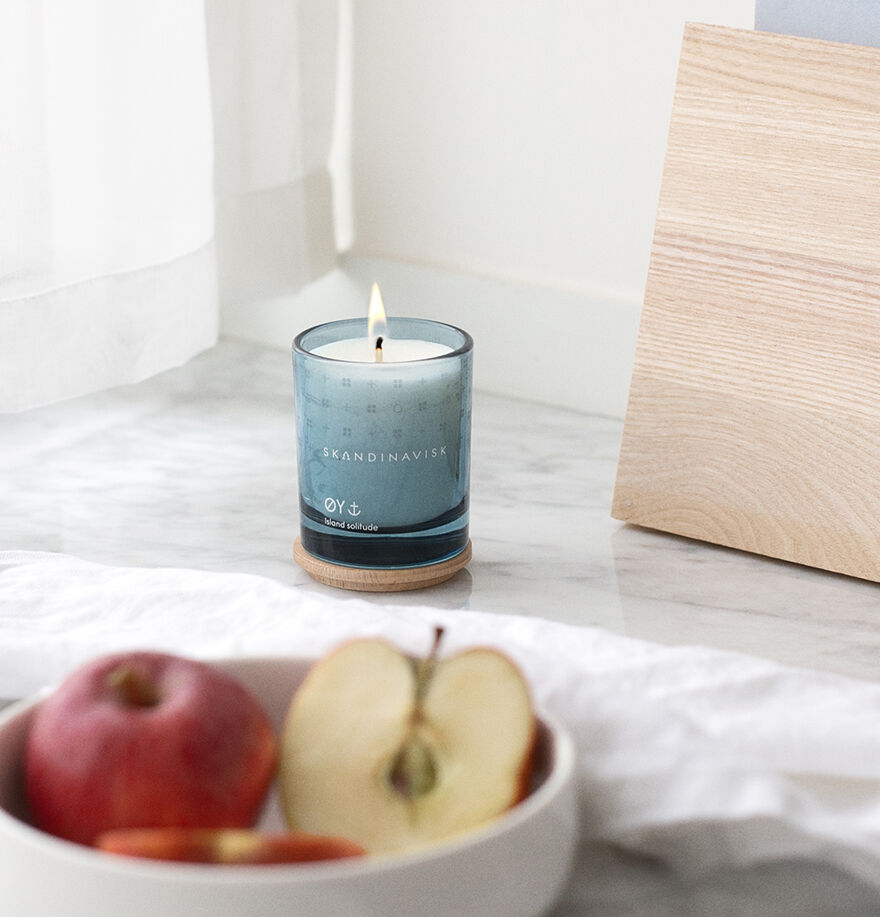 The Skandinavisk Mini Scented Candle Oy 20h, 65g, made with Swedish rapeseed wax, sits lit in its glass holder with an engraved beechwood lid on a coaster beside apples on marble near a wooden object and a white curtain.