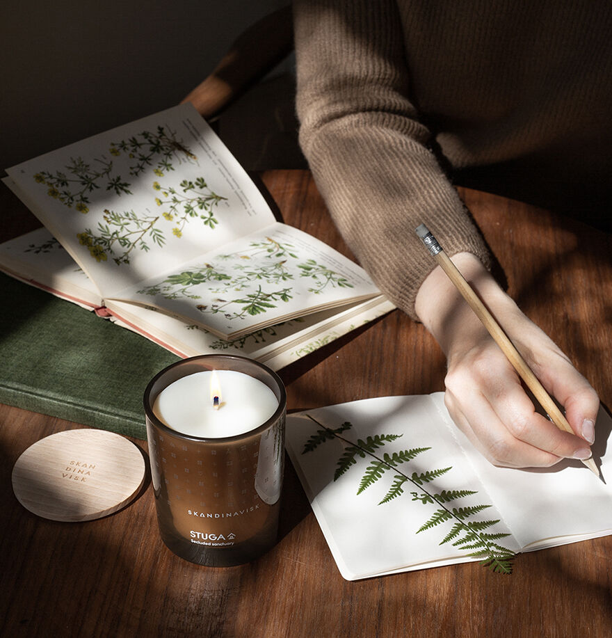 A person sketches a fern leaf in a notebook on a wooden table, beside an open botanical book, a green journal, and a lit Skandinavisk Seasonal Stuga Scented Candle 50h, 200g made from rapeseed wax.
