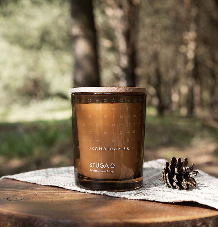 A Skandinavisk Seasonal Stuga Scented Candle 50h, 200g, sits on a wooden surface beside a pine cone, evoking woodland mosses with a blurred forest background.