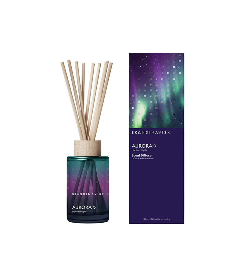 A Skandinavisk Scent Diffuser Aurora (Small) 100 ml with wooden reeds sits beside its dark purple box, adorned with a green and purple northern lights design. The alcohol-free fragrance combines soothing notes of sandalwood and juniper berries, inspired by the magical auroras that illuminate Nordic skies.