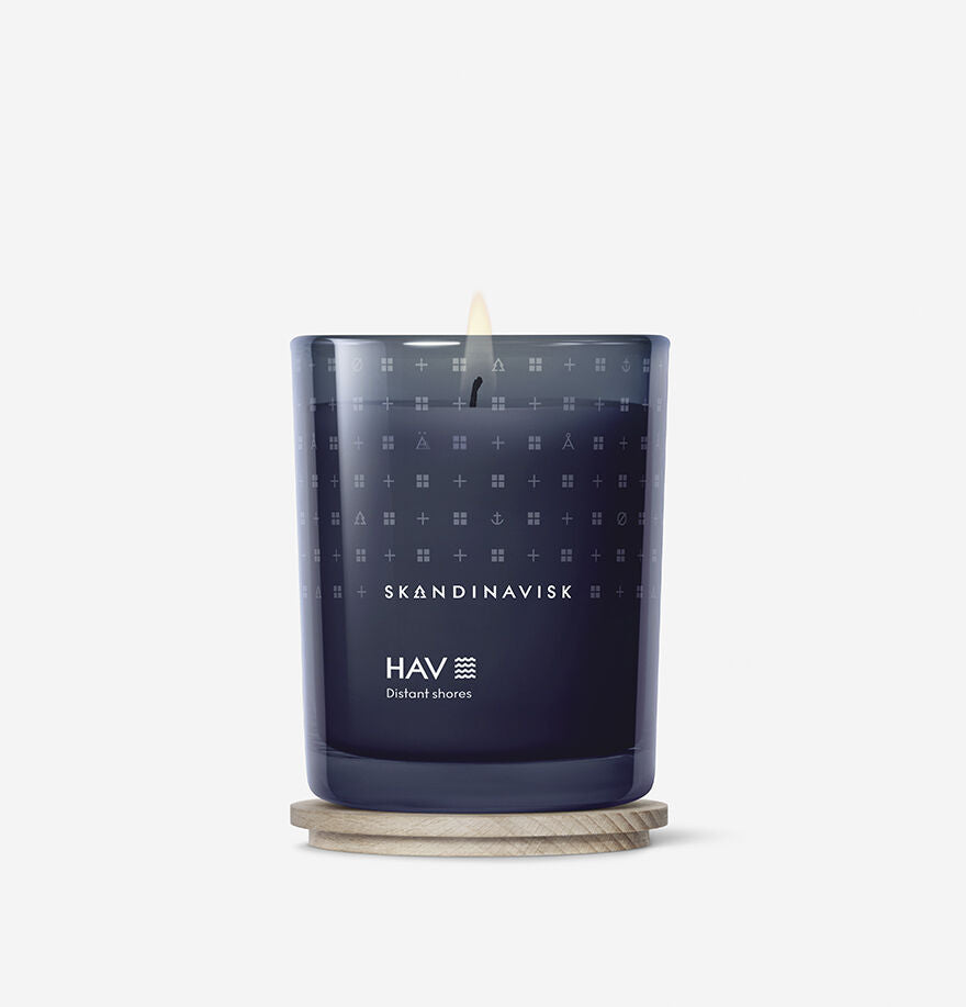 A Skandinavisk Scented Candle Hav 50h, 200g in dark blue glass, made from Swedish rapeseed wax, burns on a round wooden coaster, filling the air with the fresh scent of the Scandinavian sea against a plain white background.
