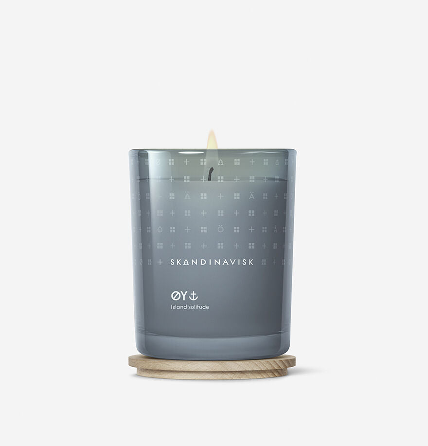 A lit Skandinavisk Scented Candle Øy, 200g, in a glass holder sits on a round wooden coaster against a plain white background.