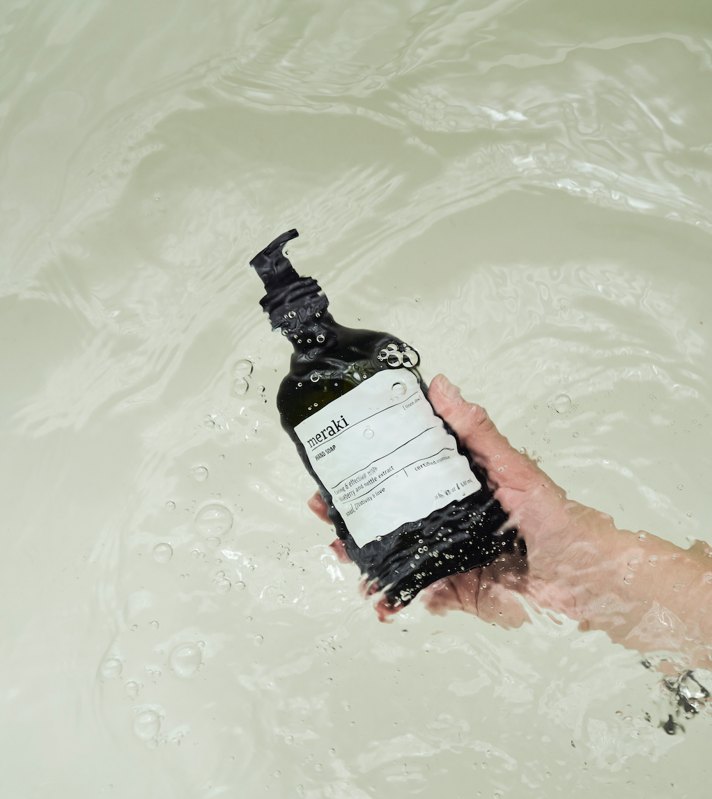 A hand holds a dark pump bottle of Meraki hand soap partially submerged in clear, rippling water.