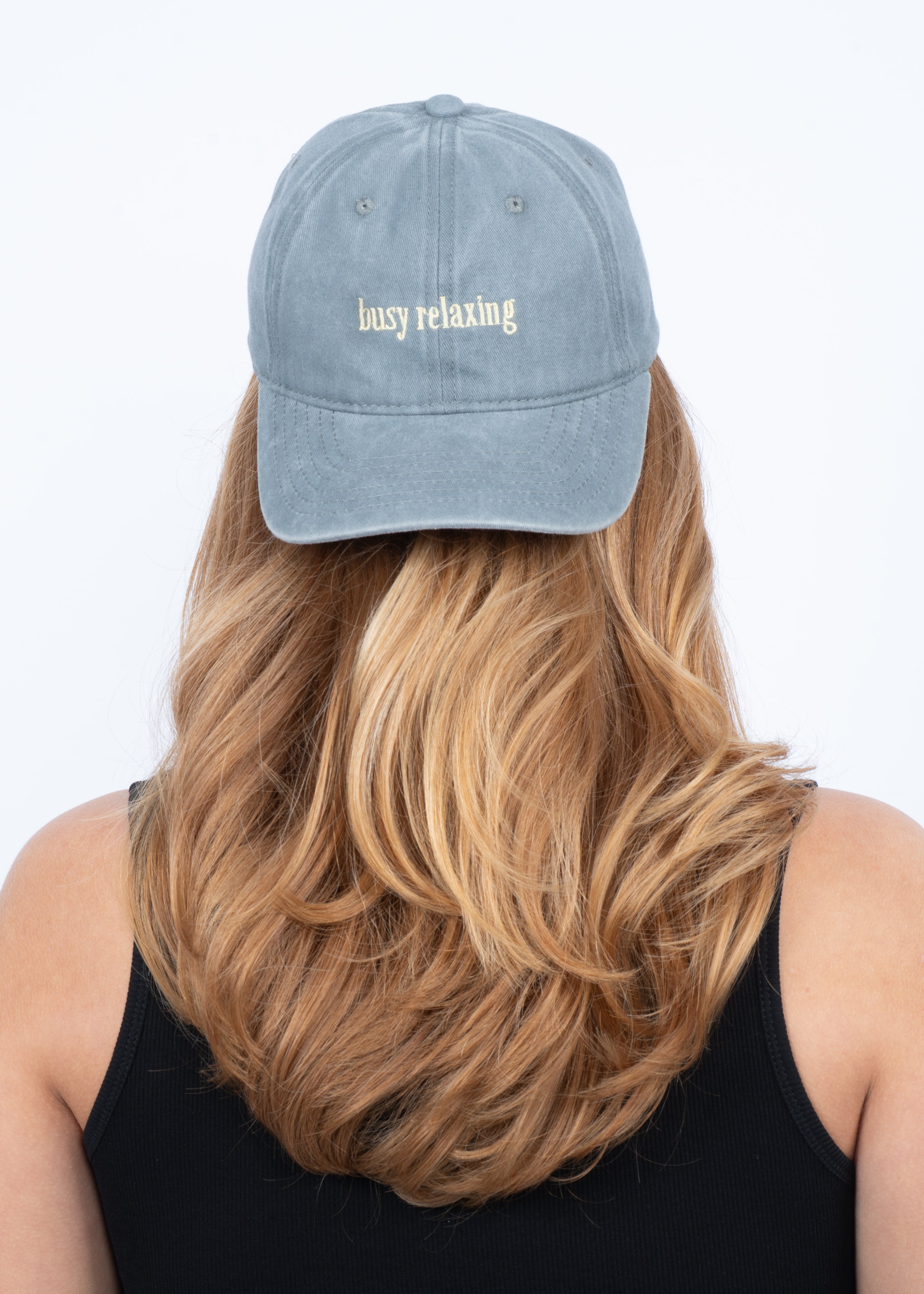 A person with long, wavy blonde hair is seen from behind wearing the Sui Ava Soft Served Cap in Busy Relaxing (Liqourice Grey) and a black sleeveless top. The cap features "busy relaxing" embroidered in white.