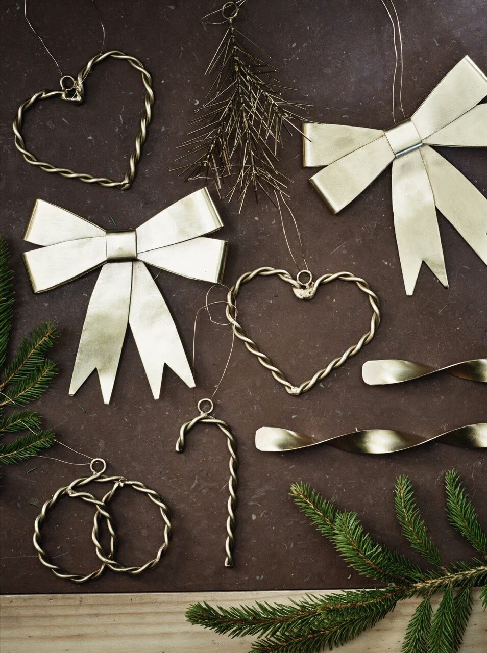 Strömshaga’s Ribbon Hanging Ornaments in brass, featuring elegant shapes like bows, hearts, a candy cane, circles, and a branch, are beautifully displayed on a brown surface with green pine sprigs in the corners—creating the perfect brass ornament collection to decorate your Christmas tree or accentuate a festive ribbon display.