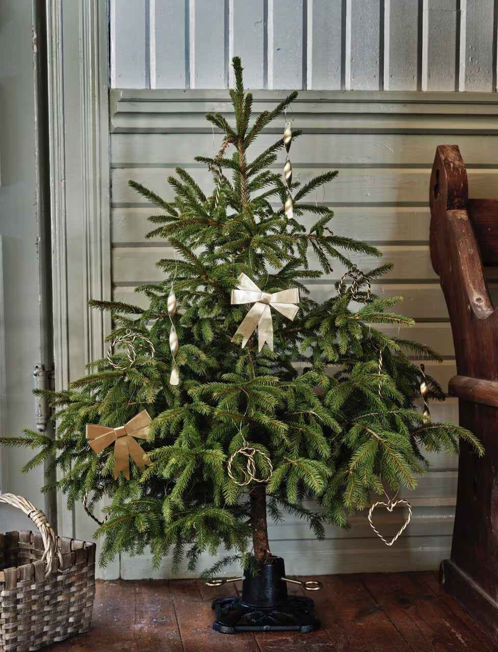 A small Christmas tree adorned with Strömshaga’s brass Ribbon Hanging Ornaments, along with gold bows, spiral ornaments, decorative ribbon, and heart-shaped decorations, stands on a wood floor near a basket and a wooden bench against a paneled wall.