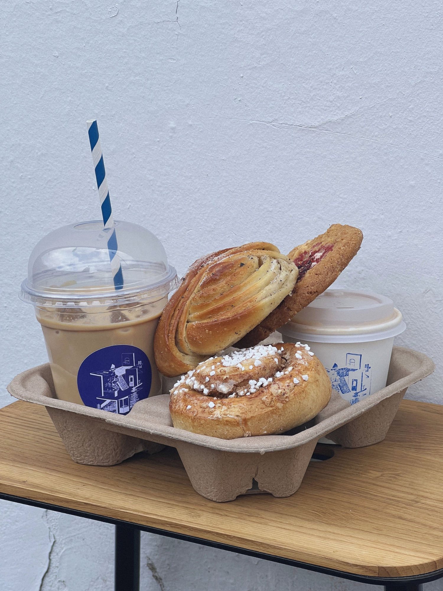 Cardboard tray with pastries and a coffee drink on a wooden surface against a light gray wall.