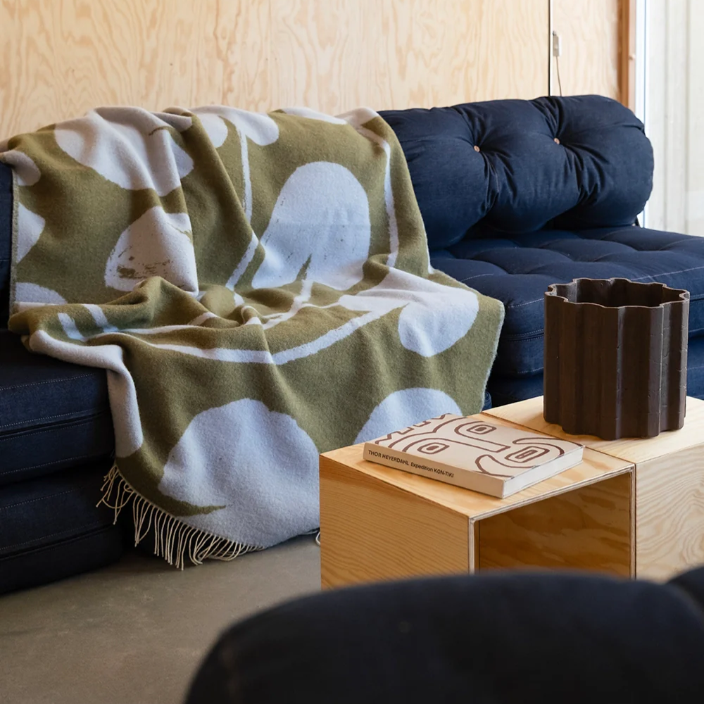 A cozy living room features a dark blue tufted sofa draped with a green and white patterned blanket. In front, a light wood table holds a wavy-edged dark vase and a book with a bold, abstract face design on the cover.