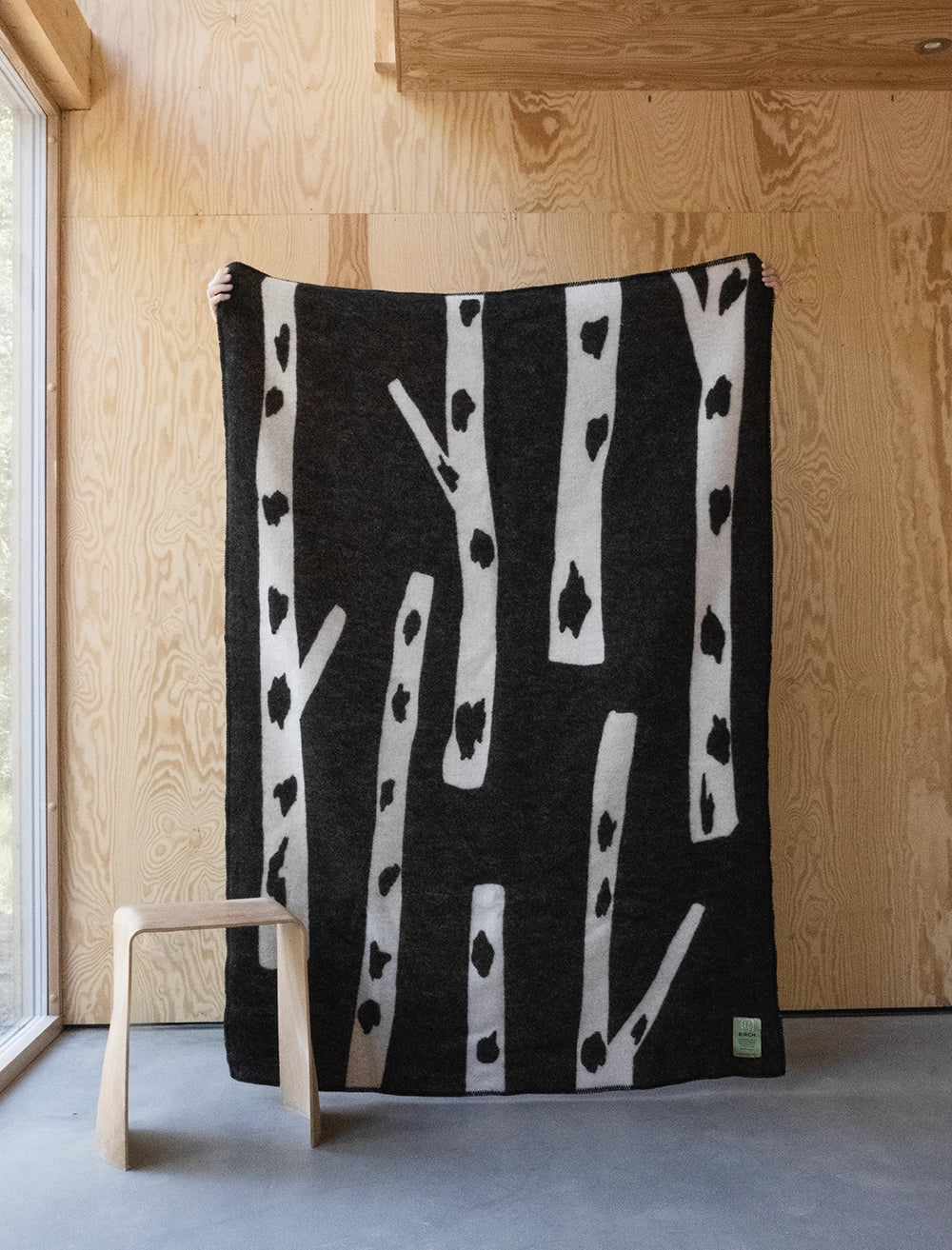 A person holds the Fine Little Day Birch Wool Blanket, Reversible in Black & White, standing in a wooden room beside a stool and large window—a scene reflecting classic Scandinavian design.