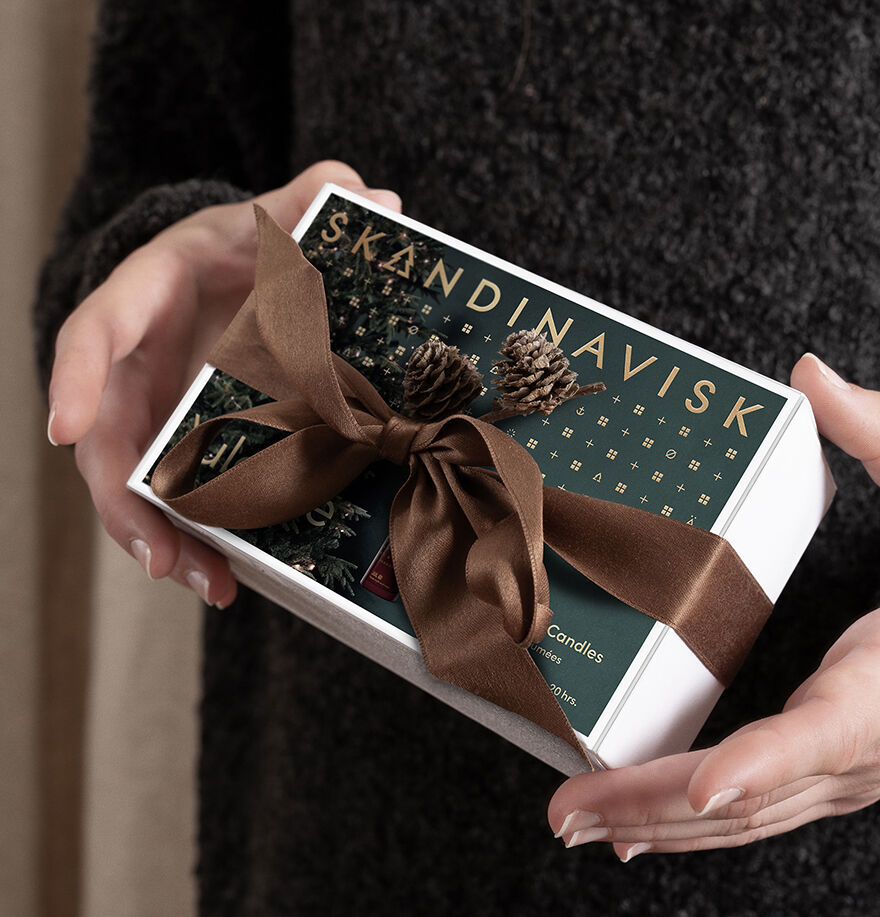 A person holding the Skandinavisk Giftset Yuletide—featuring two scented candles, Jul & Skog—wrapped with a brown ribbon and pinecones, making it the perfect seasonal gift for candle lovers and mini scented candle enthusiasts.