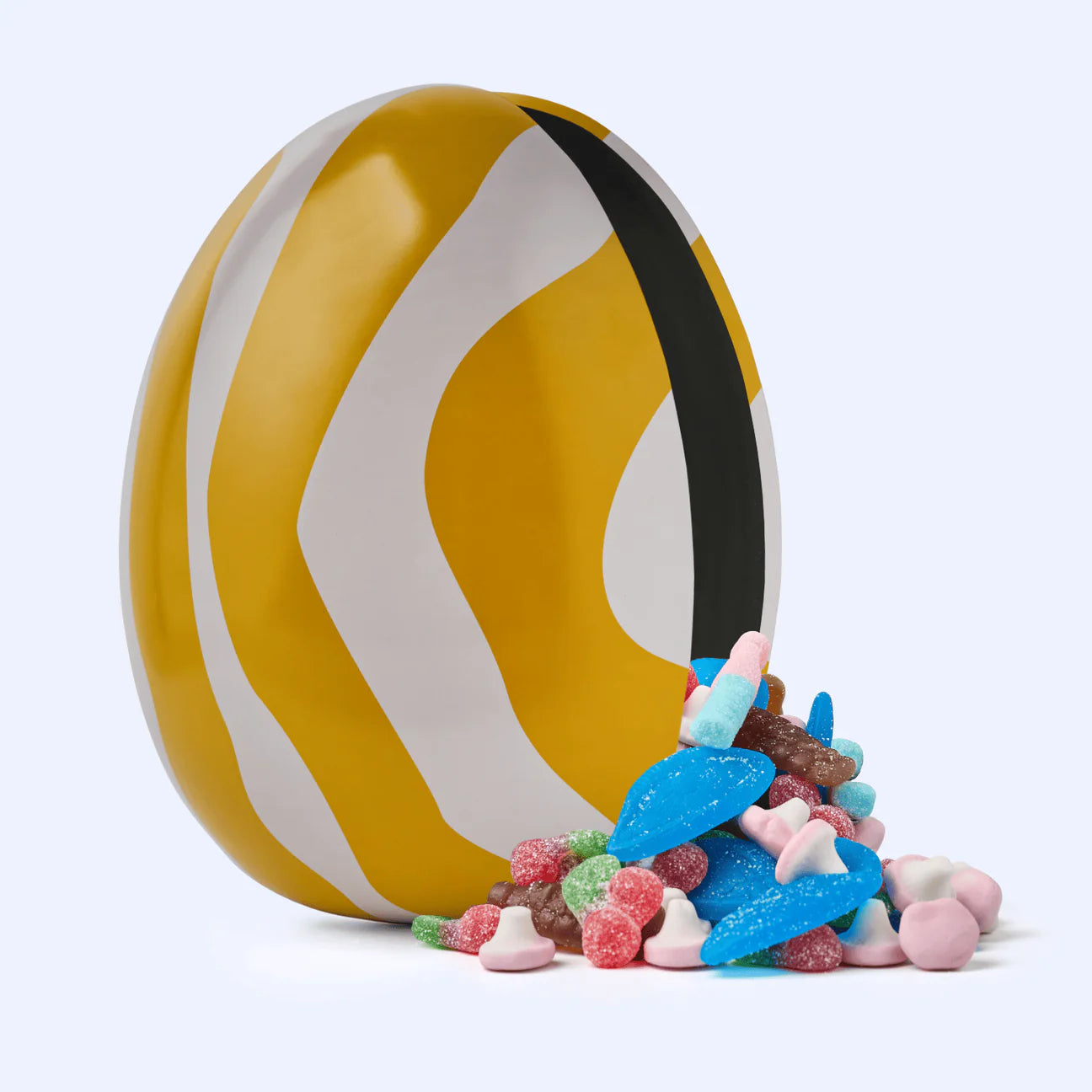The Gåva Gifting Zebran Big Reusable Easter Egg, a Swedish-style tin Påskägg with yellow, white, and black wavy stripes, is tilted on its side as colorful assorted candy spills out.