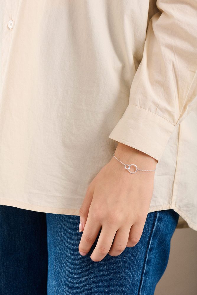 Wearing a cream long-sleeve shirt and blue jeans, a person showcases Pernille Corydon’s Double Bracelet in Silver, featuring two circles for a delicate yet striking look on their wrist.