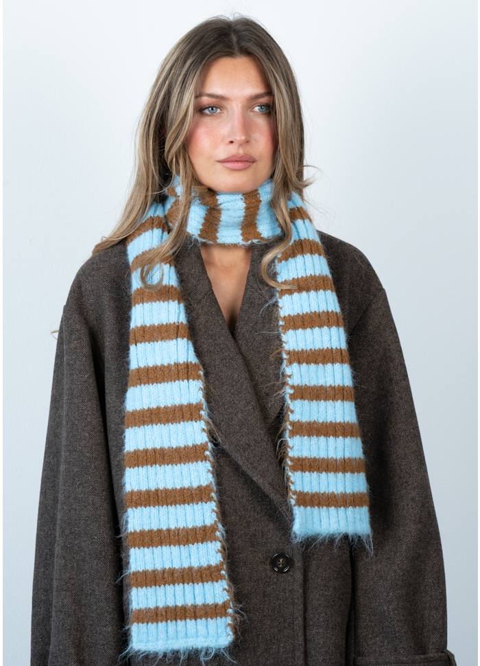 A person wearing a dark brown coat and the Clara Slim Scarf in Blue and Khaki from Sui Ava, standing against a plain light background.