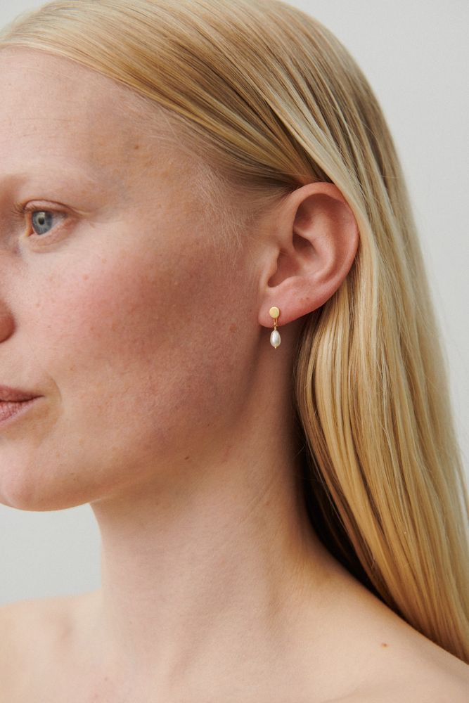 A close-up of a person with long, straight blonde hair wearing Pernille Corydon’s Ocean Dream Earrings in Gold with freshwater pearls. A neutral background highlights the elegance of these signature earrings.