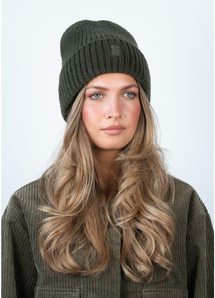 A woman with long wavy blonde hair wears the Sui Ava Brenda Beanie in Dark Green and a matching corduroy jacket, looking straight ahead with a neutral expression against a plain light background.