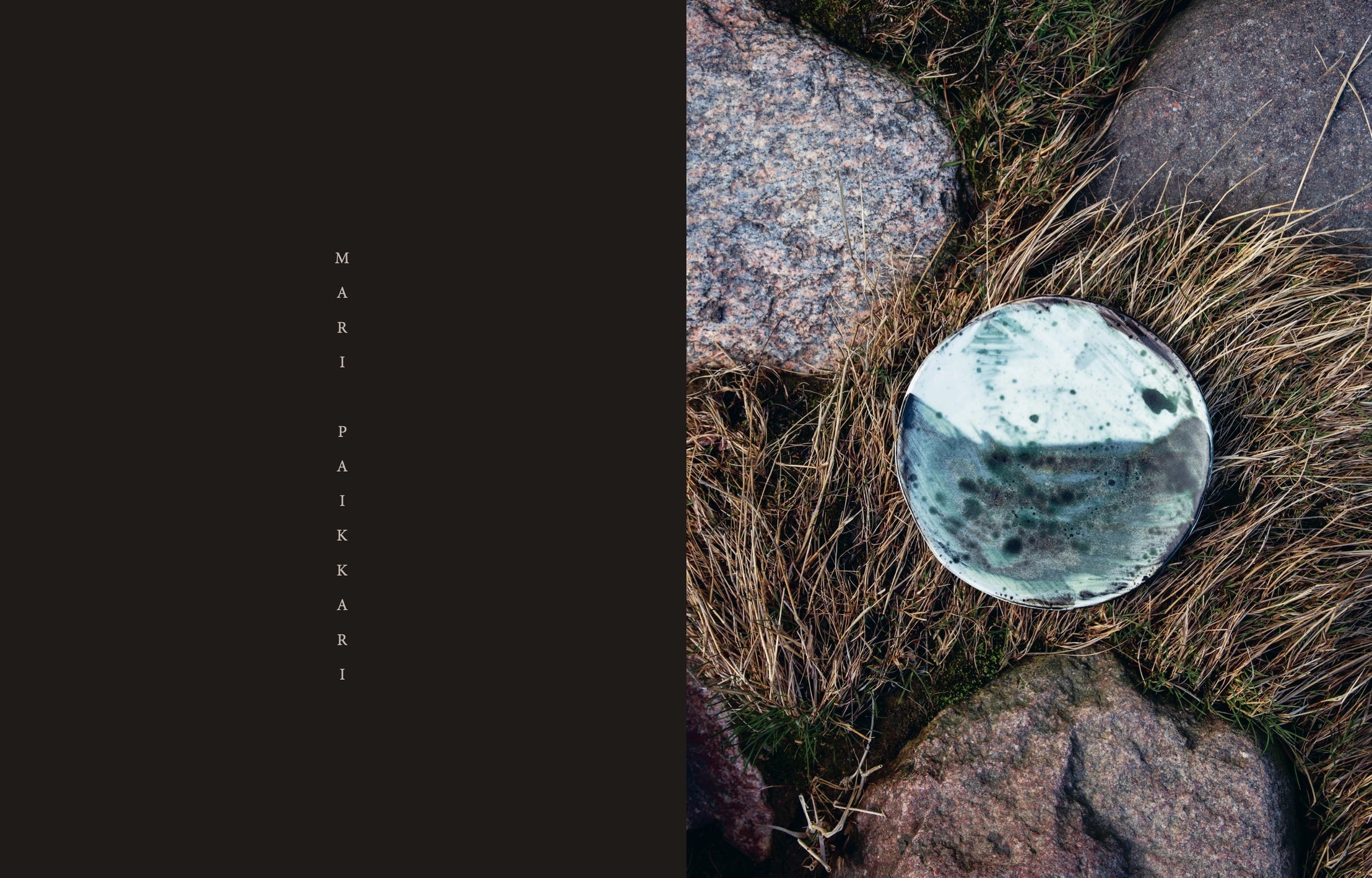 A book titled "Genuine – Handmade Beauty from the North" by Cozy Publishing rests on dry grass among rocks. To the left, "MARI PAIKKARI" is written vertically in spaced-out letters on a black background.