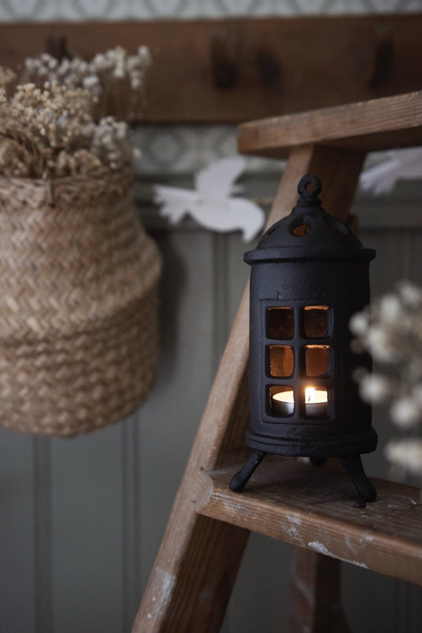 A cozy scene features the Strömshaga Stove Candle Holder in black cast iron with a lit candle on a wooden ladder, accompanied by a hanging basket of dried flowers and white paper decorations.