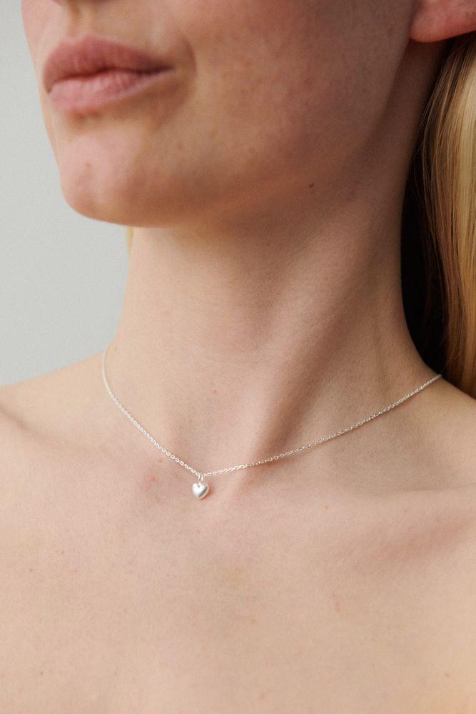 A close-up of a woman wearing the Pernille Corydon Love Necklace in Gold, featuring a delicate heart-shaped pendant on a fine chain, set against a plain light background with her shoulders and neck visible.