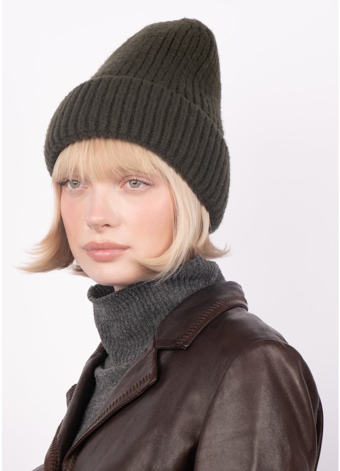 A woman with short blonde hair wears the Brenda Beanie in Dark Green by Sui Ava, paired with a gray turtleneck sweater and a brown leather jacket, looking slightly to the side against a plain light background.