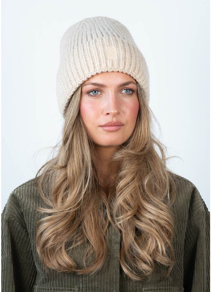 A woman with long, wavy blonde hair wears the Sui Ava Brenda Beanie in Beige and an olive corduroy jacket, looking straight ahead with a neutral expression against a plain light background.