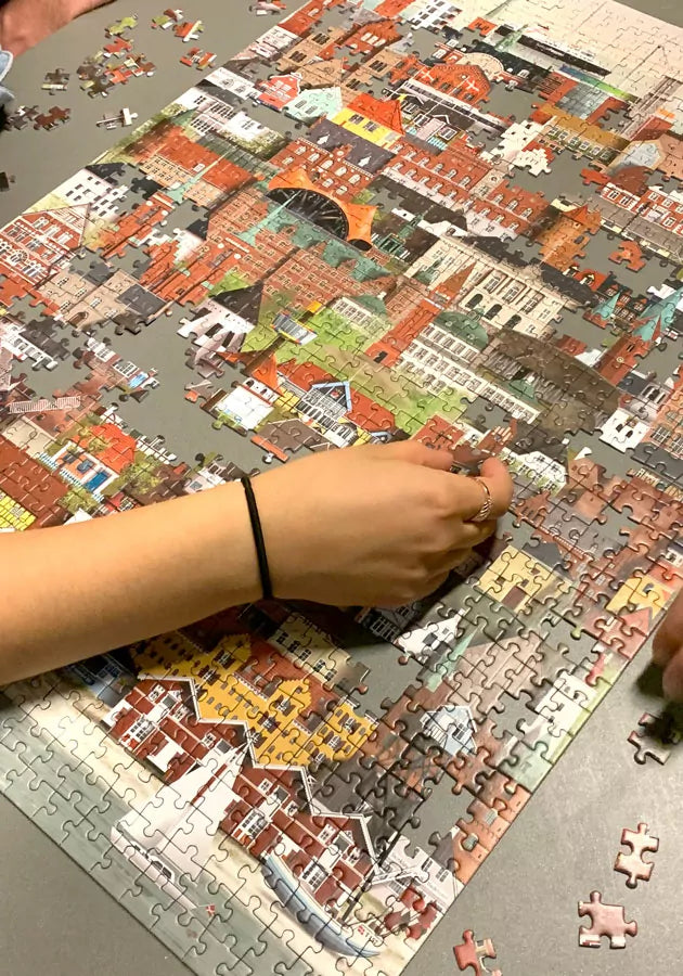 Two people are assembling the Martin Schwartz Denmark Puzzle, 1000 Pieces, featuring a colorful cityscape on a grey table. One person’s hand holds a piece in the foreground while pieces are scattered around the edges.