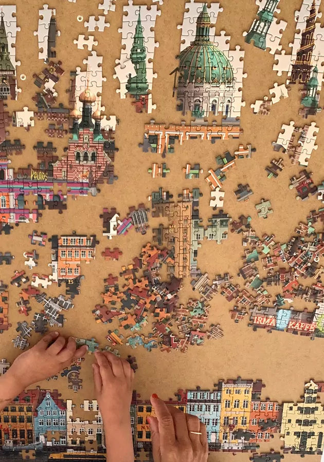 Multiple hands collaborate to assemble the Martin Schwartz Copenhagen City Puzzle, 1000 Pieces, featuring vibrant buildings and city landmarks. Numerous pieces from this colorful puzzle remain scattered on the wooden surface, yet to find their place.