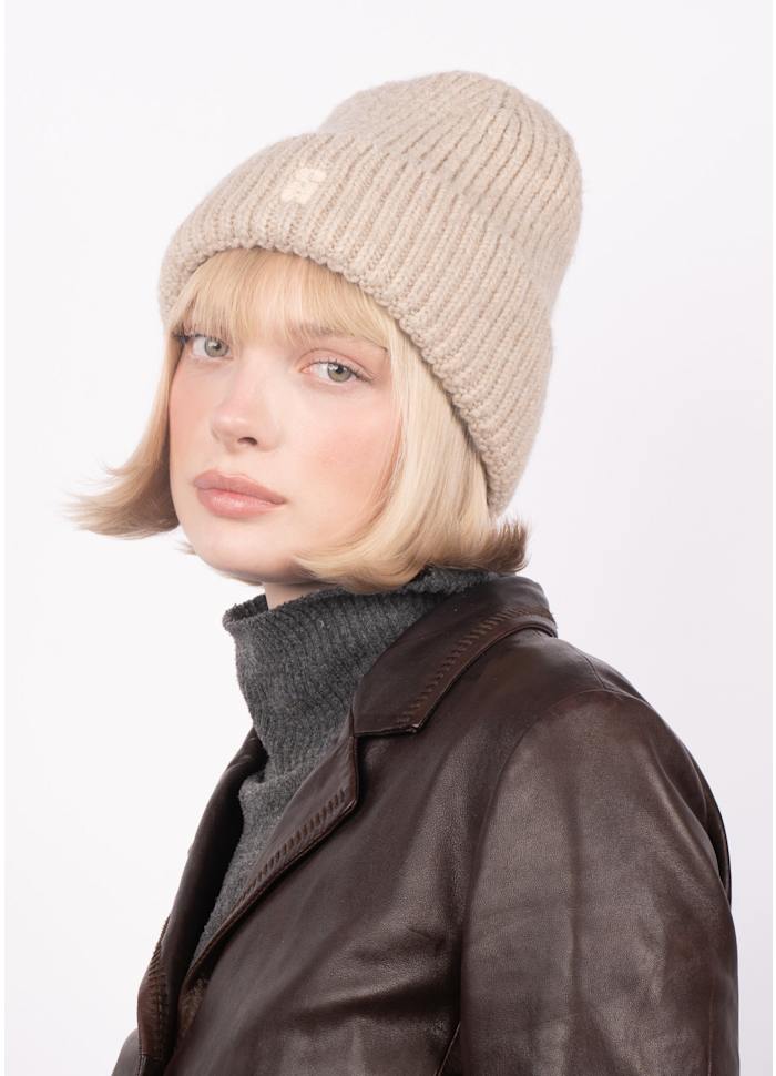 A woman with chin-length blonde hair wears the Brenda Beanie in Beige by Sui Ava, paired with a dark brown leather jacket and a gray turtleneck. She looks slightly to the side against a plain light background.