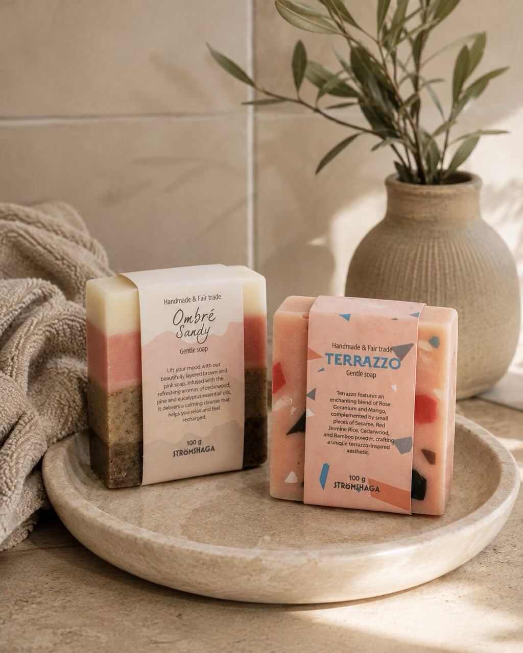 Two artisanal soap bars—Strömshaga’s Terrazzo Handmade & Fairtrade Soap Bar and the Ombre Sandy bar—crafted from handmade vegan soap, rest on a beige dish beside a towel. A small vase and plant in the background complete the natural, spa-like scene.