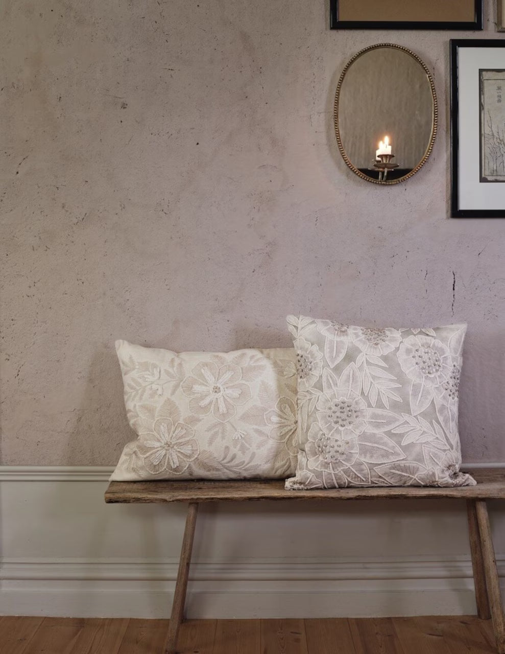 Two Strömshaga Cushion Covers Roslinna (40x60 cm) in embroidered beige floral design rest on a wooden bench beneath an oval mirror with a lit candle and framed art, creating a cozy, decorative setting.